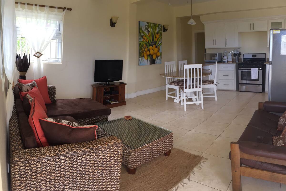 A living room with a sofa, a coffee table, and a television. There is a dining area with a table and chairs. The kitchen is visible with appliances and cabinets.
