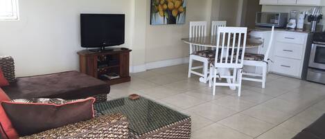 A living room with a brown sofa, a patterned ottoman, a TV on a wooden stand, and a painting of coconuts on the wall.