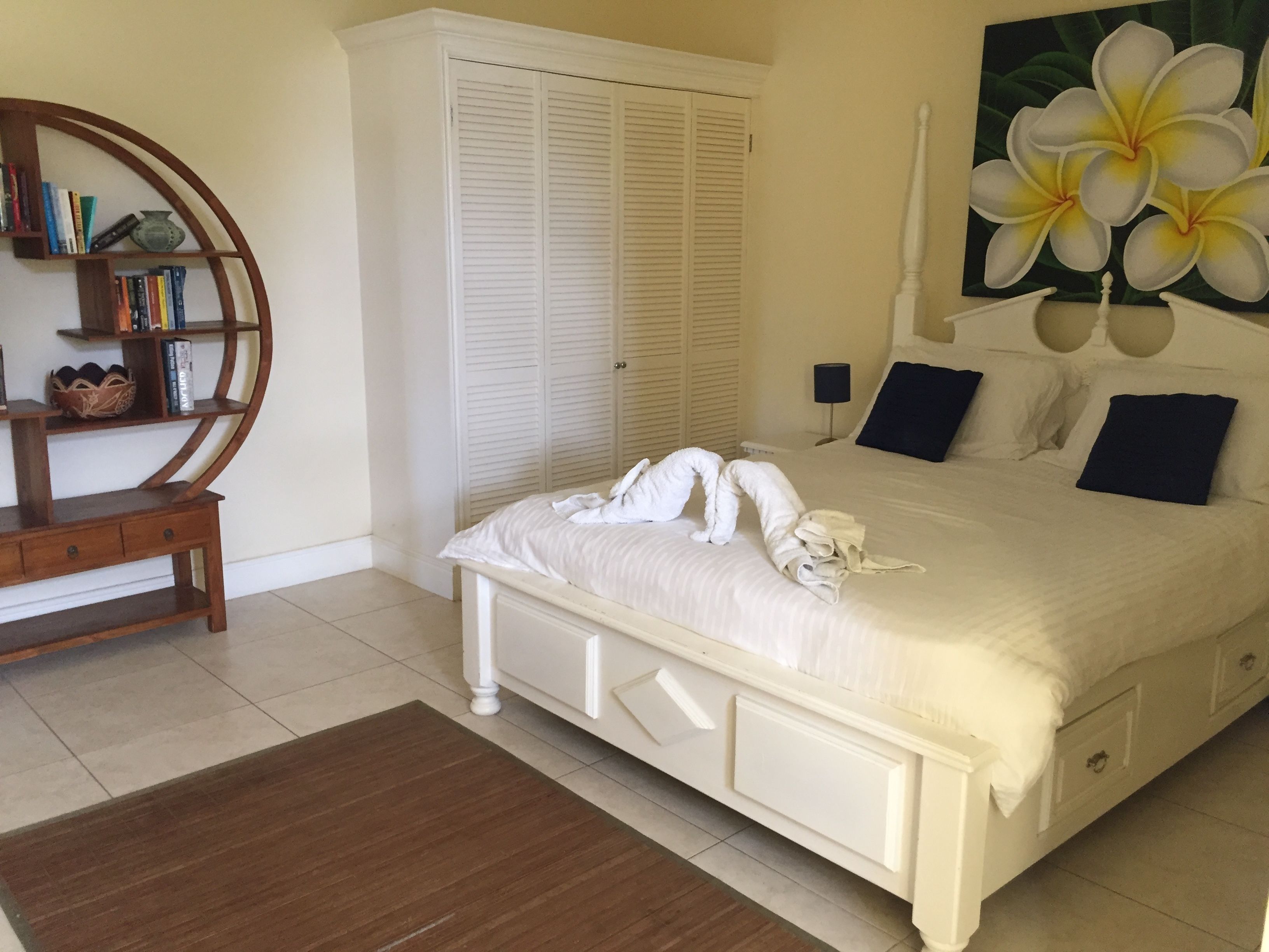 A neatly made bed with towels, a wooden side table, a bookshelf, and a large painting of flowers on the wall.