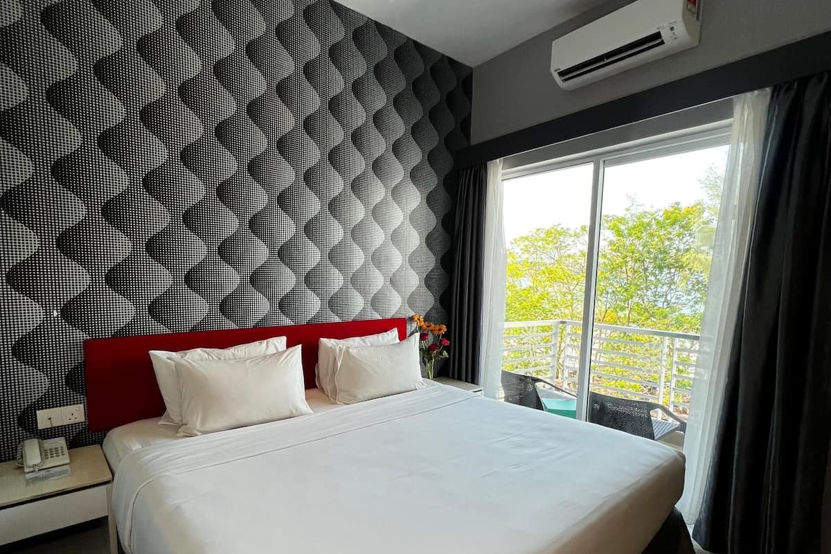 Una habitación de hotel moderna con una cama grande, un cabecero rojo, aire acondicionado empotrado en la pared y una ventana con vistas a zonas verdes.