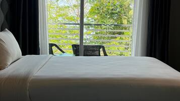 A hotel room with a large bed, a view of greenery and trees through a window, and a chair outside the window.