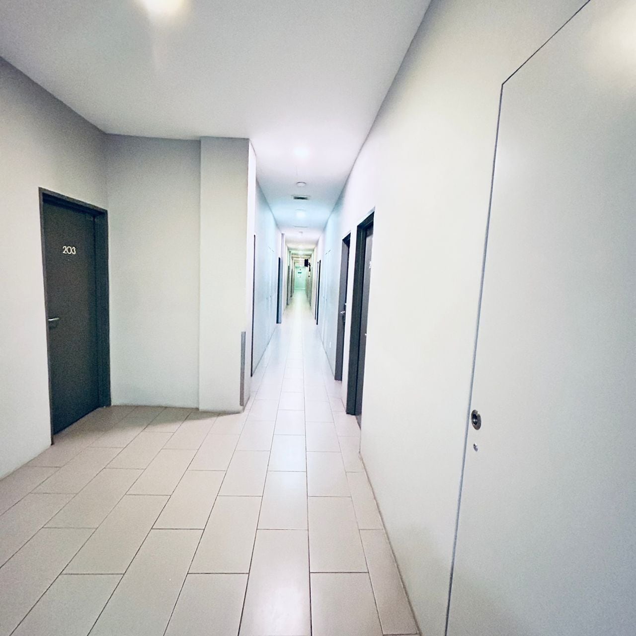 A long, narrow hallway with white tiled floors and walls, featuring doors on both sides.