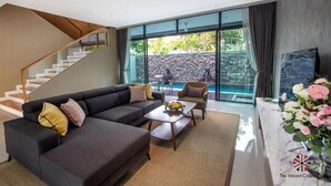 Una sala de estar moderna con una escalera, un conjunto de sofás, una mesa de centro y vistas a una piscina y un muro de piedra.