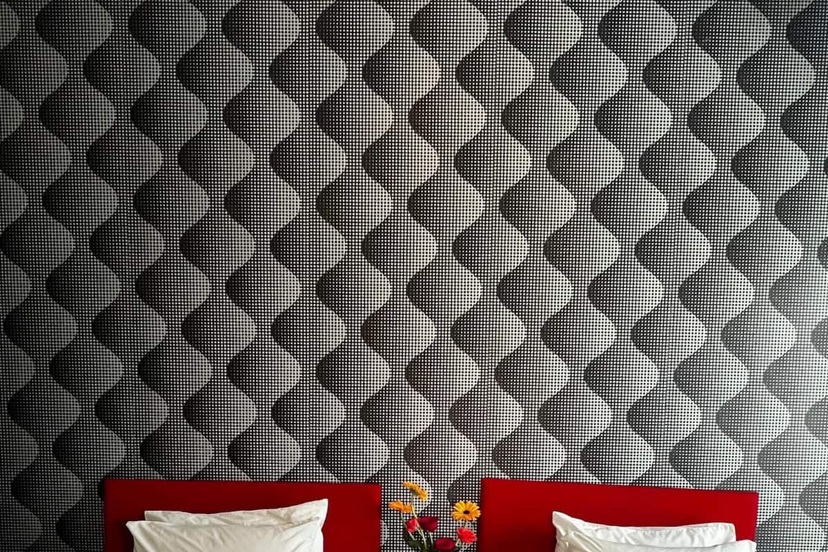 Habitación de hotel con dos camas, un cabecero rojo, una mesita con frutero y flores, y una pared con diseño.