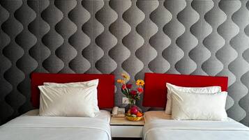 A hotel room with two beds, a red headboard, a small table with a fruit bowl and flowers, and a patterned wall.