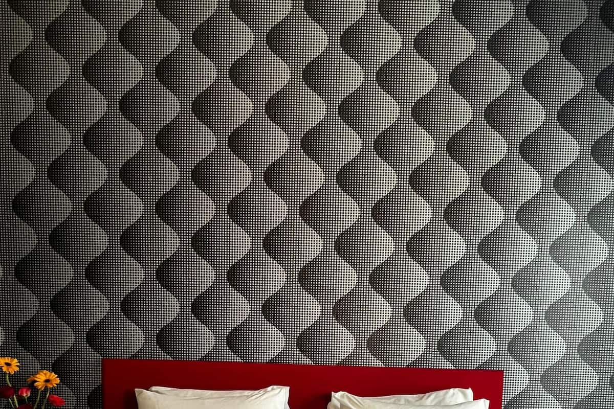 Habitación de hotel con cabecera roja, dos almohadas blancas, cubrecama blanco, un florero con flores y un frutero sobre la mesita de noche.