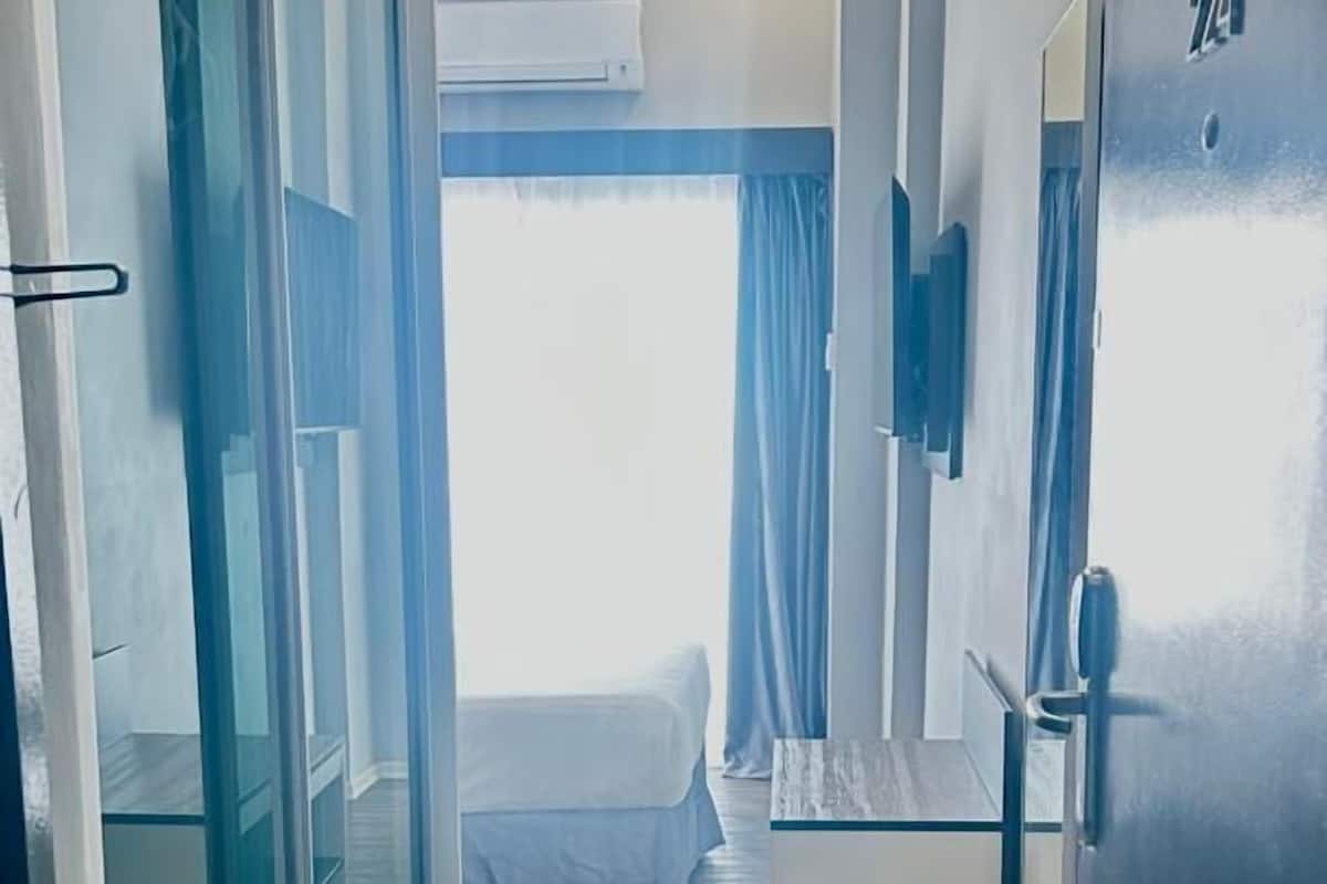 Pasillo de hotel con una puerta corredera de vidrio, una cama a lo lejos y una puerta con placa de identificación.