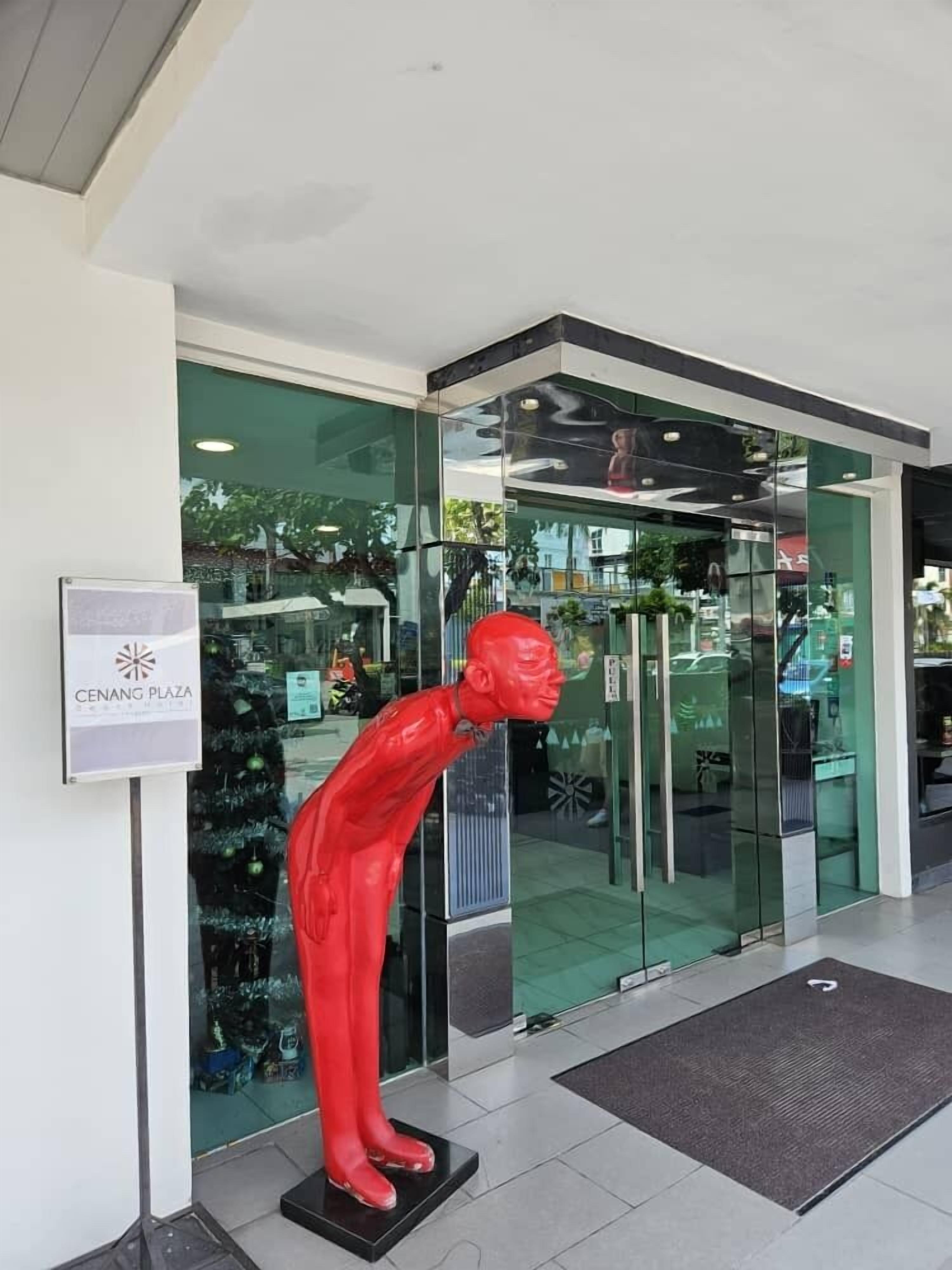 A modern building entrance with a glass facade, a red statue in front, and a sign for Cenang Plaza.