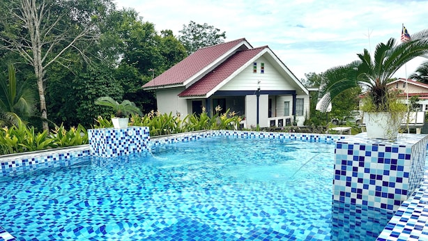 Una piscina con azulejos de mosaico azul, rodeada de abundante vegetación y una casa con tejado rojo.