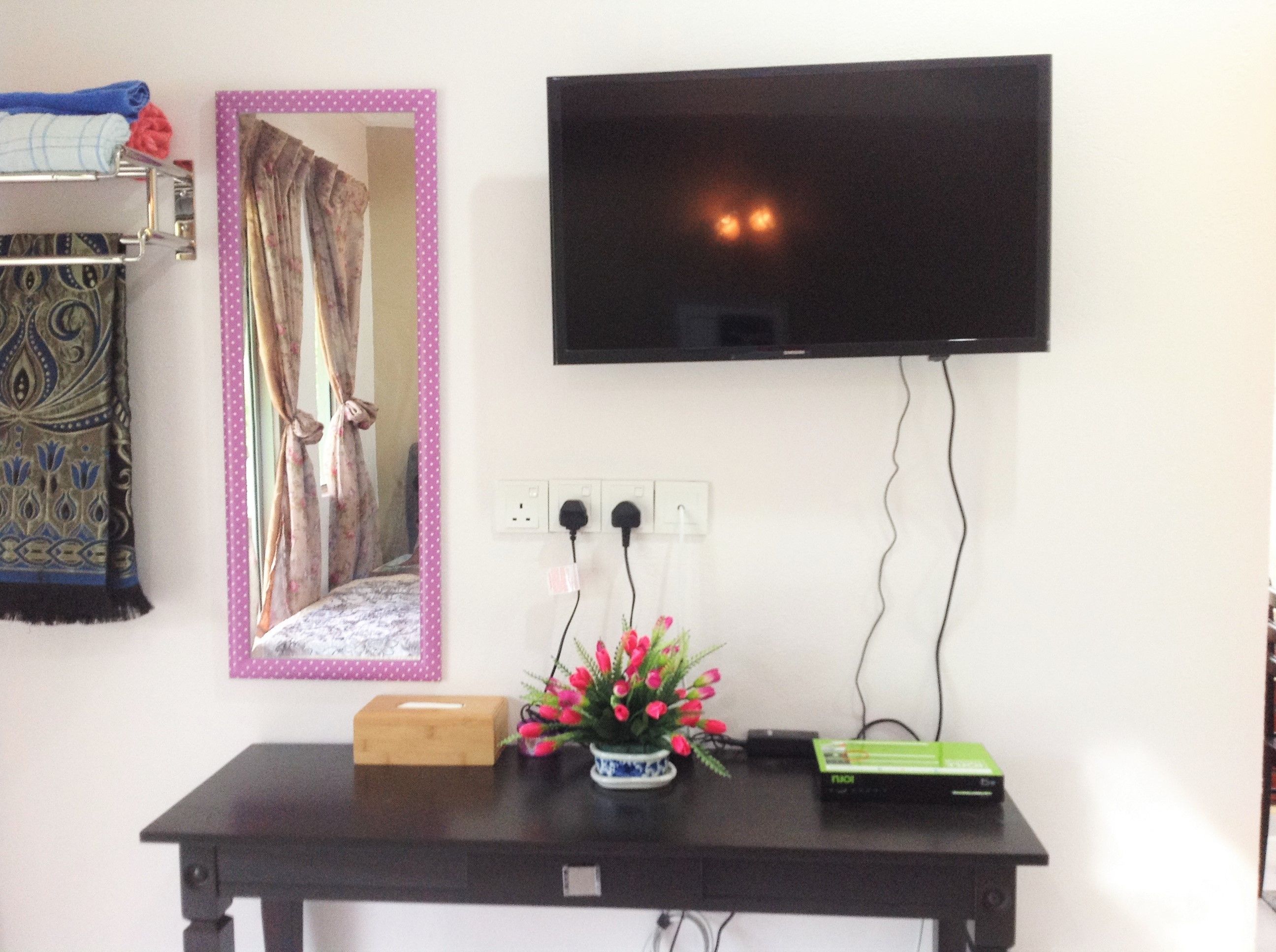 Una habitación con una televisión montada en la pared, un espejo con marco rosa, una mesa con una caja y un florero con flores, y un enchufe.