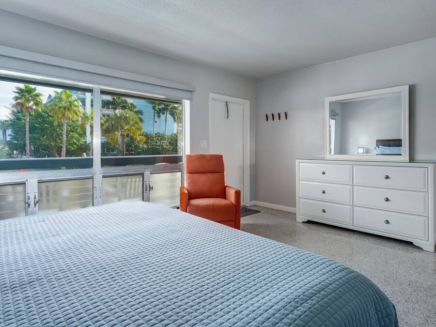 A bedroom with a large bed, a white dresser, an orange chair, and a large window with a view of palm trees.