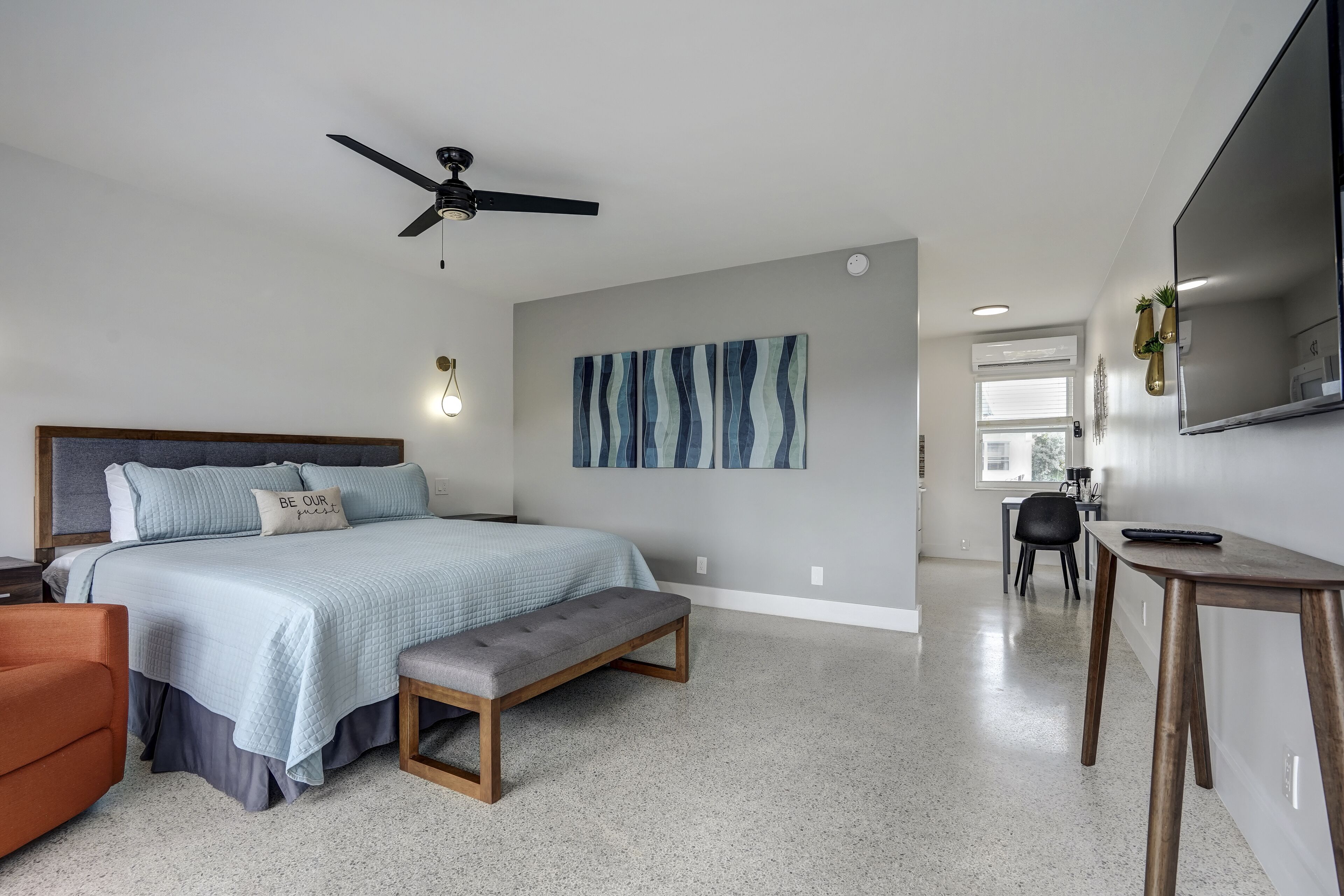 Un dormitorio moderno con cama, banca, ventilador de techo, obra de arte y una zona de comedor con mesa y sillas.