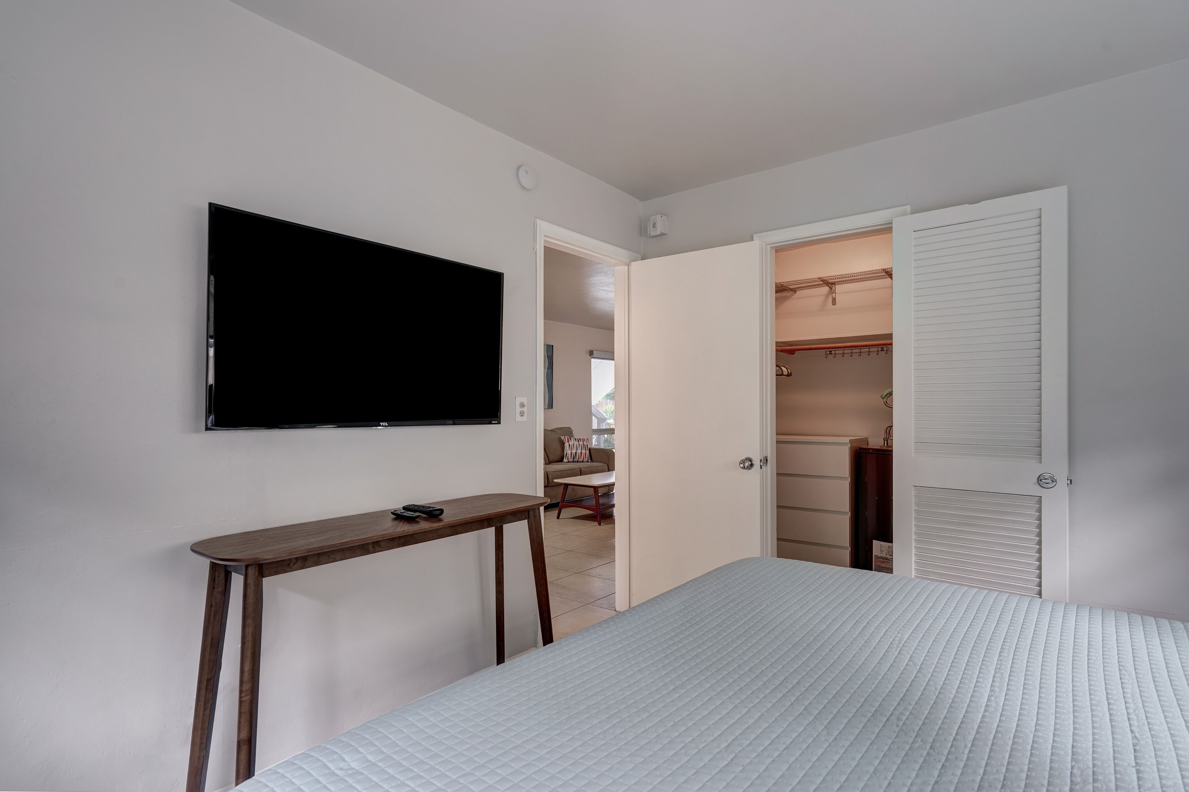 A bedroom with a bed, a TV mounted on the wall, a wooden side table, and an open closet.