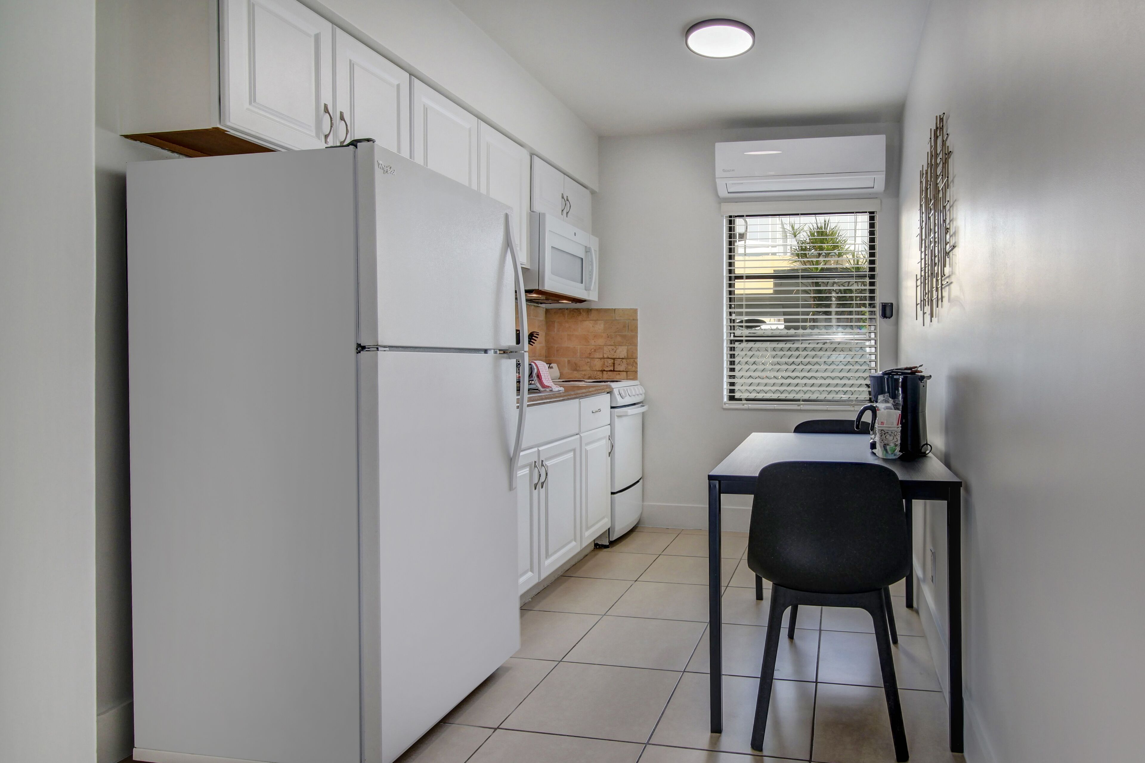 Cocina compacta con armarios blancos, un refrigerador blanco, un microondas y una pequeña mesa con una silla.