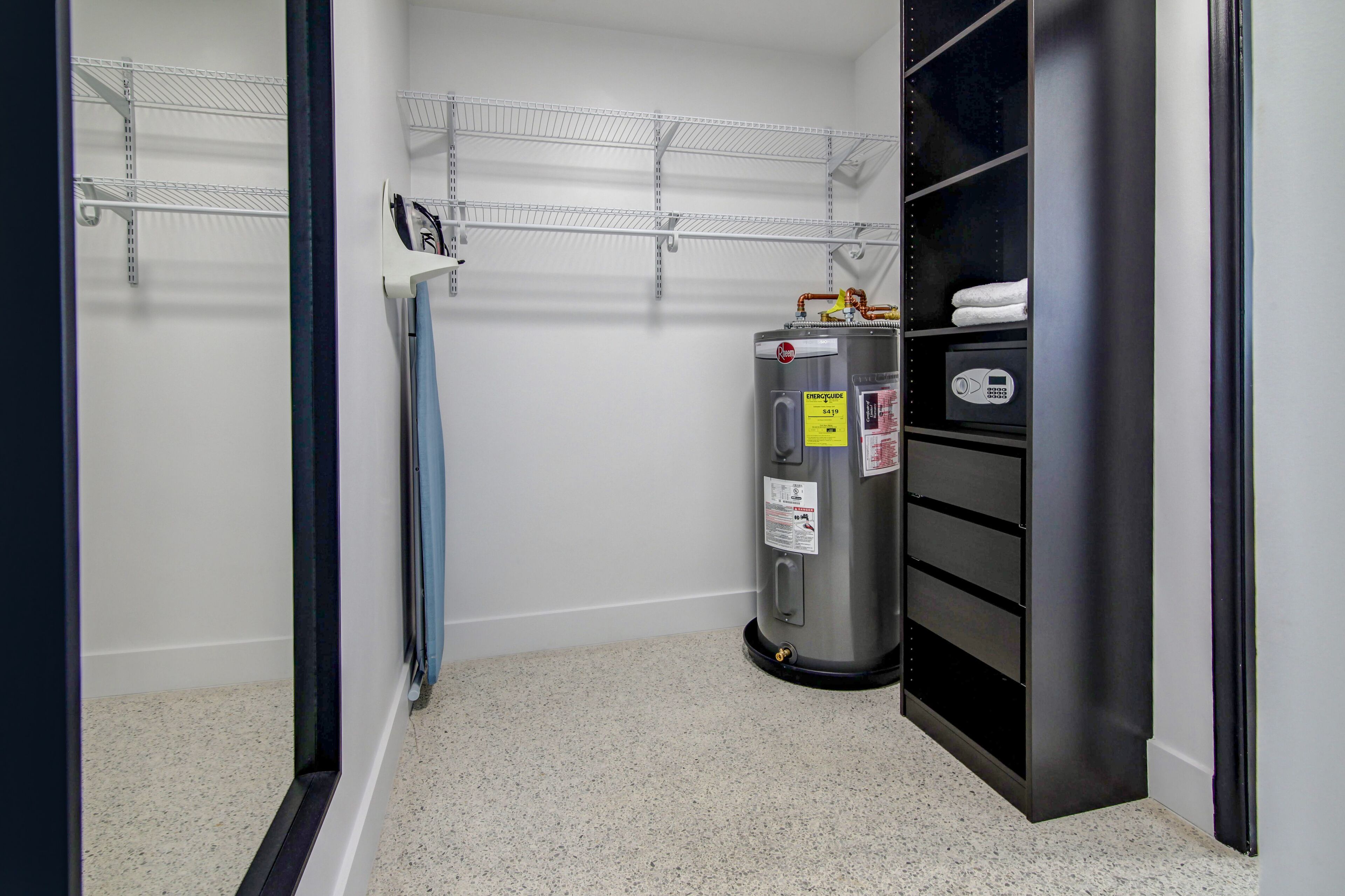 Rincón de una habitación de hotel con un estante metálico, un calentador de agua y un mueble de almacenamiento negro con perchero.