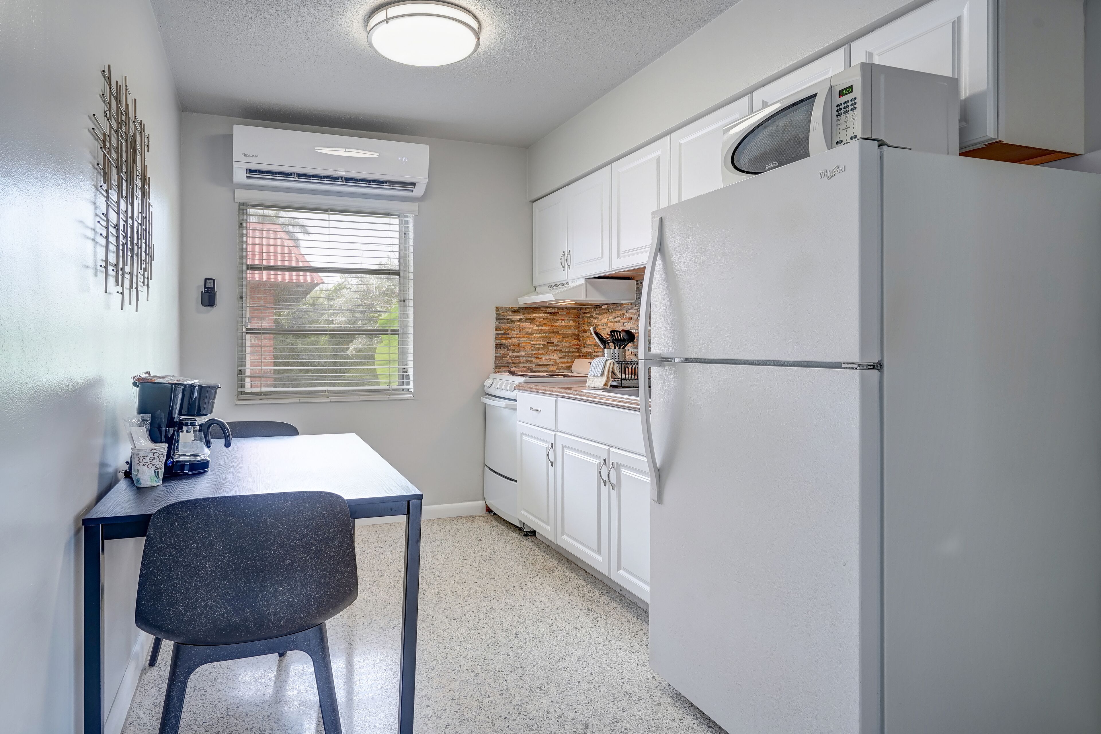 Una cocina compacta con armarios blancos, un refrigerador de acero inoxidable y una ventana con persianas.