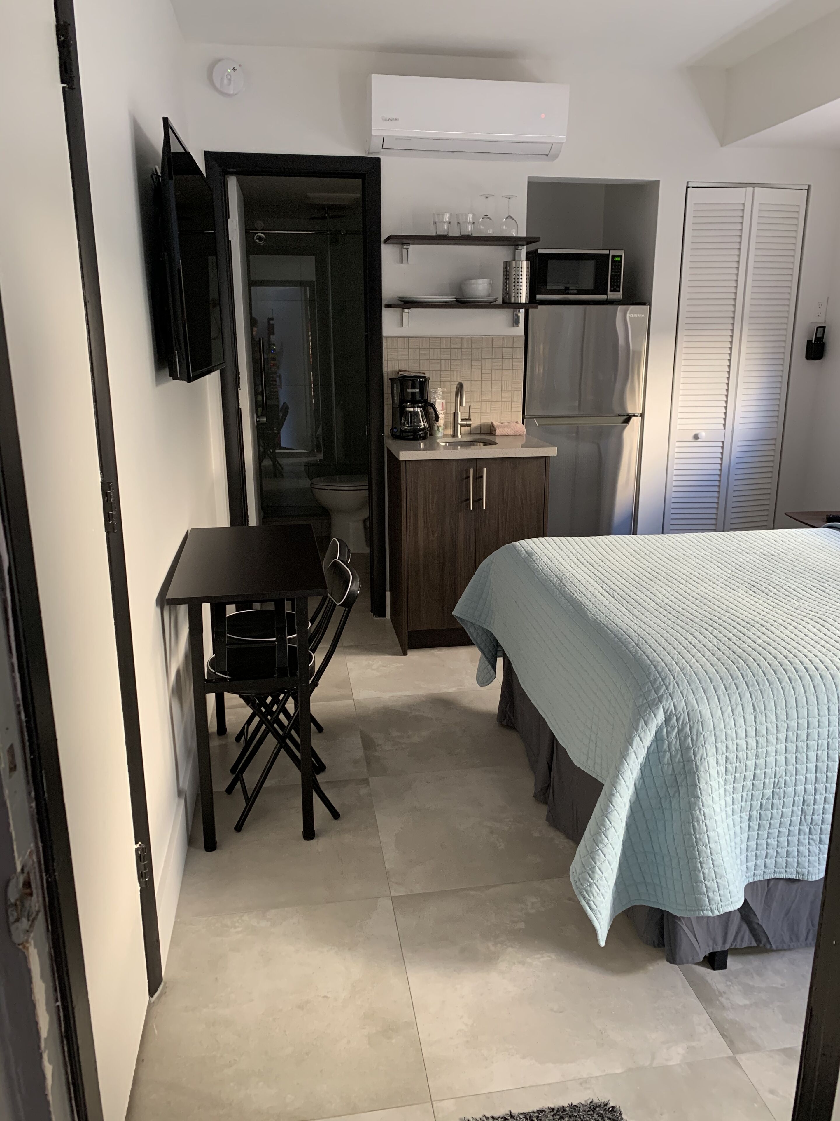 Ein gemütliches Schlafzimmer mit Bett, kleinem Tisch und Stuhl. Angrenzend an das Schlafzimmer befindet sich eine Küchenzeile mit Mikrowelle, Kaffeemaschine und Schränken.