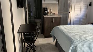 Ein gemütliches Schlafzimmer mit Bett, kleinem Tisch und Stuhl. Angrenzend an das Schlafzimmer befindet sich eine Küchenzeile mit Mikrowelle, Kaffeemaschine und Schränken.