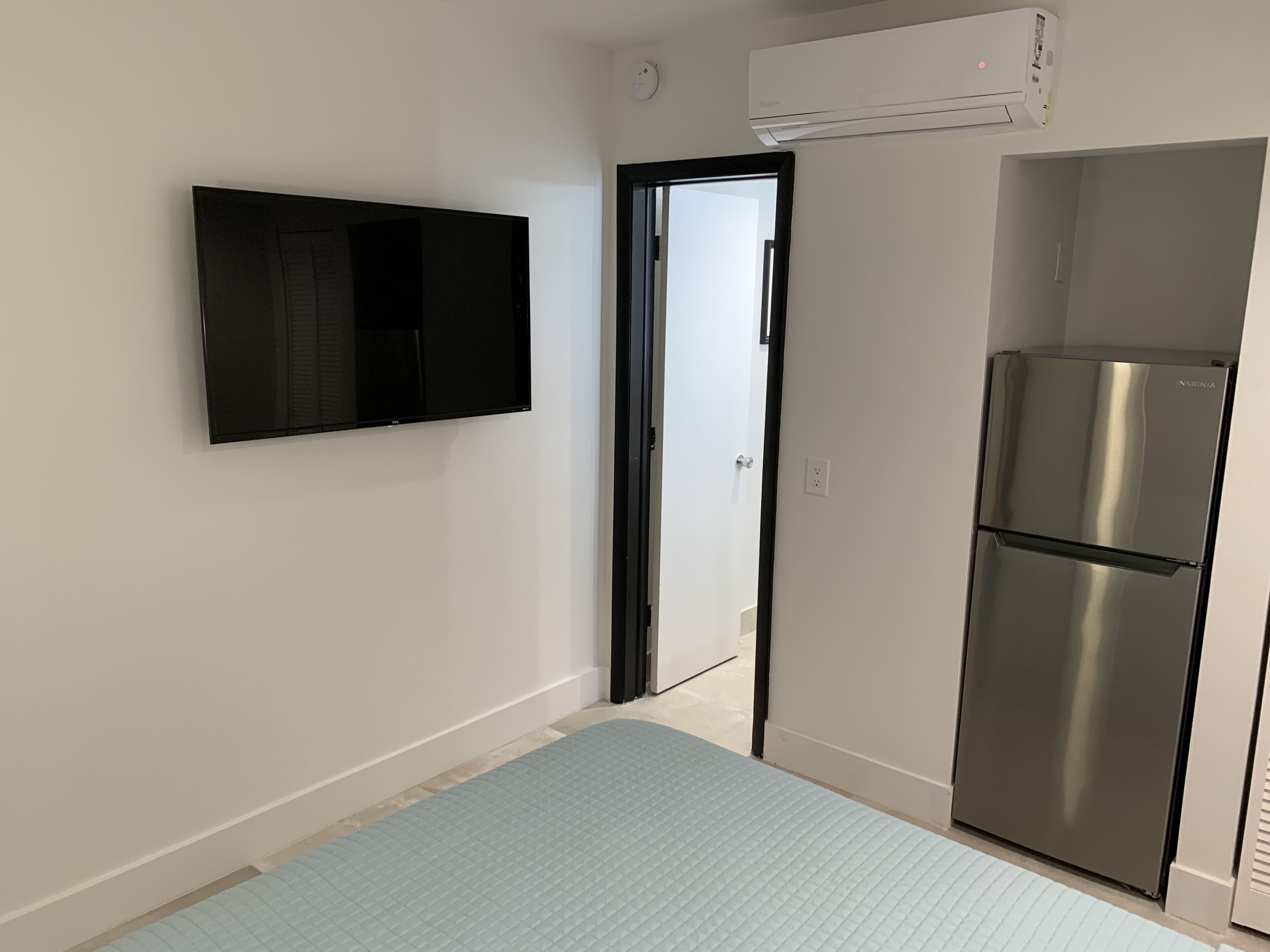 A compact room with a wall-mounted TV, a stainless steel refrigerator, and a door leading to another space.