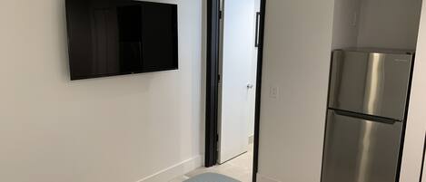 A compact room with a wall-mounted TV, a stainless steel refrigerator, and a door leading to another space.