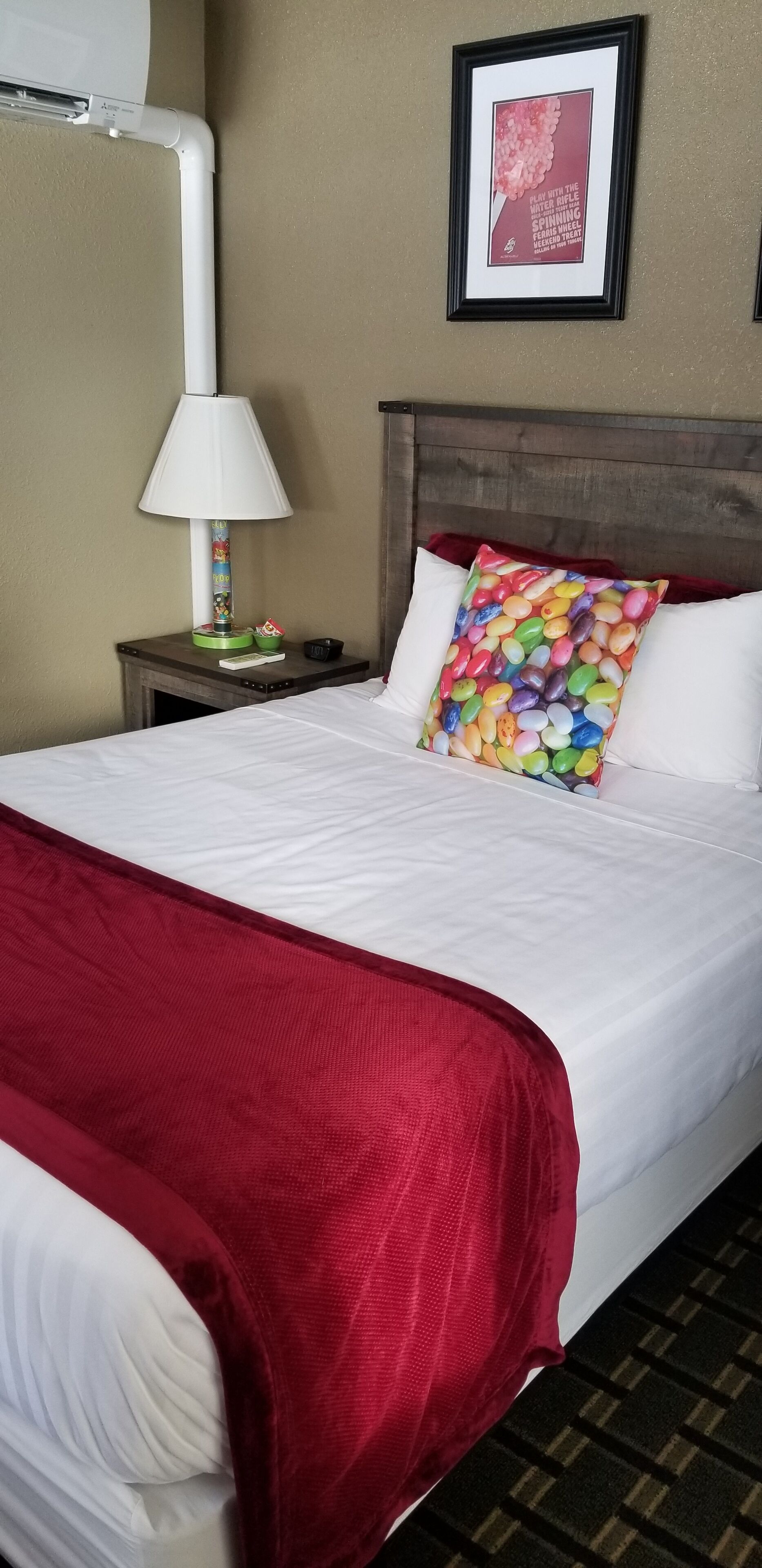 A hotel room with a neatly made bed, a bedside lamp, a framed picture on the wall, and a red throw blanket.