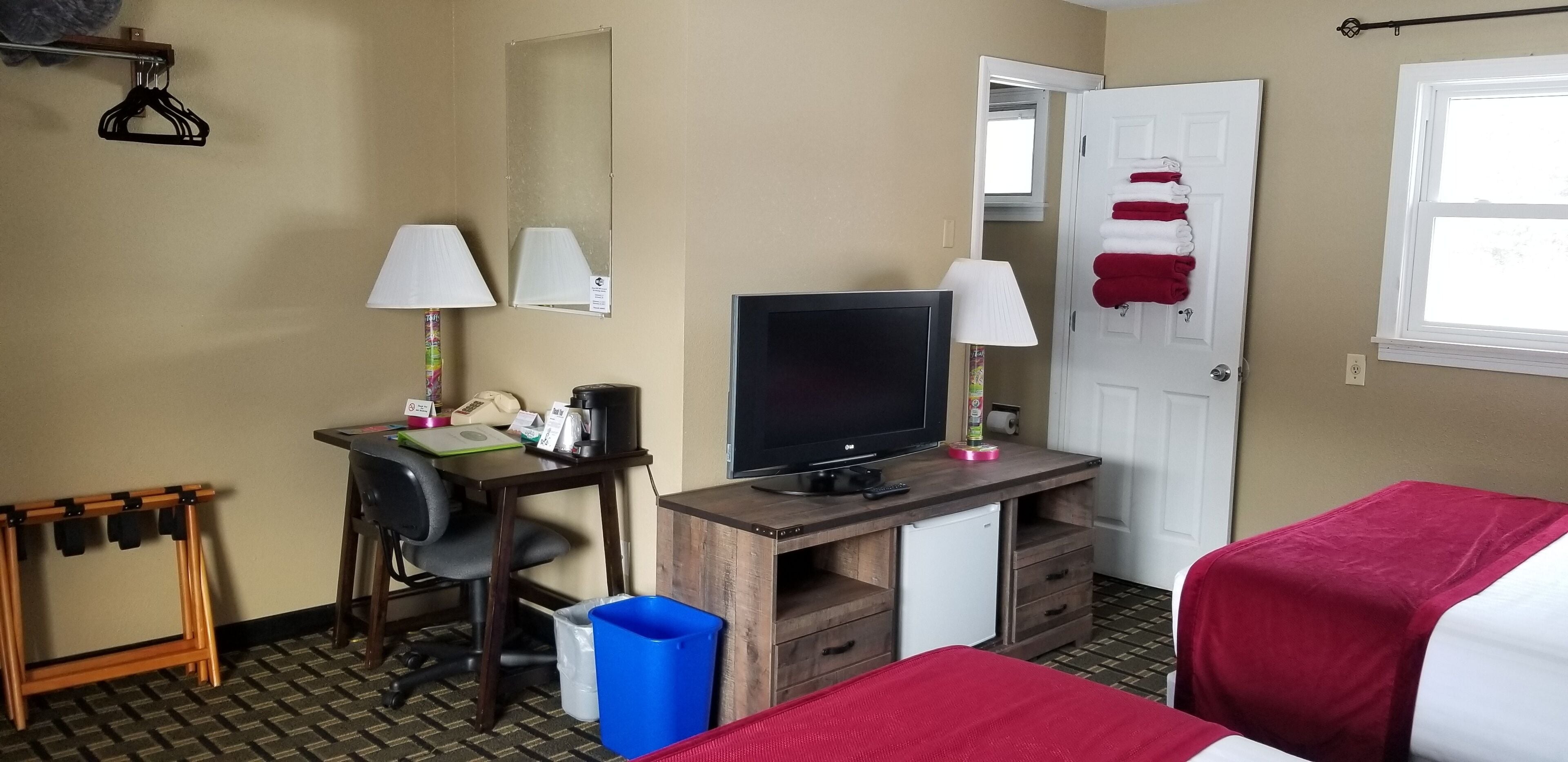 A hotel room with a desk, two lamps, a television, a mini-fridge, a trash bin, and a bed with a red blanket.