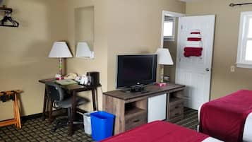 A hotel room with a desk, two lamps, a television, a mini-fridge, a trash bin, and a bed with a red blanket.