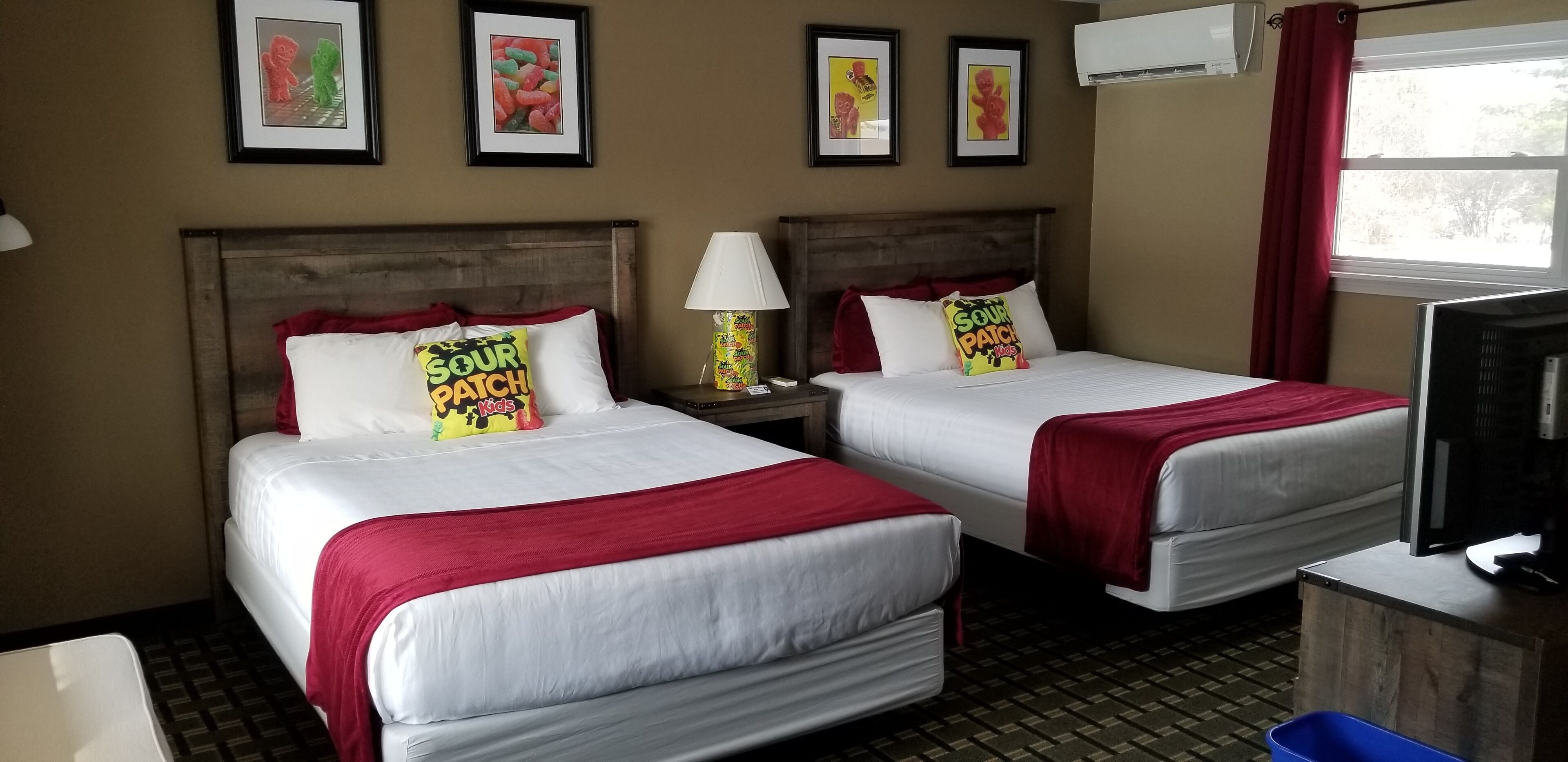 A hotel room with two beds, each with white bedding and red accents, a television on a wooden stand, and a window with red curtains.