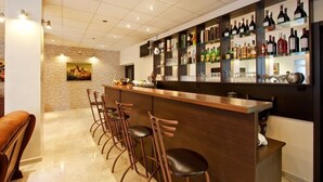 A bar area with a long counter and multiple stools, a wall of liquor bottles, and a stone wall with a painting.