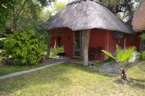 A thatched-roof house with a red exterior, surrounded by greenery and a small path leading to the entrance. - Jump Street Chalets (Maun)