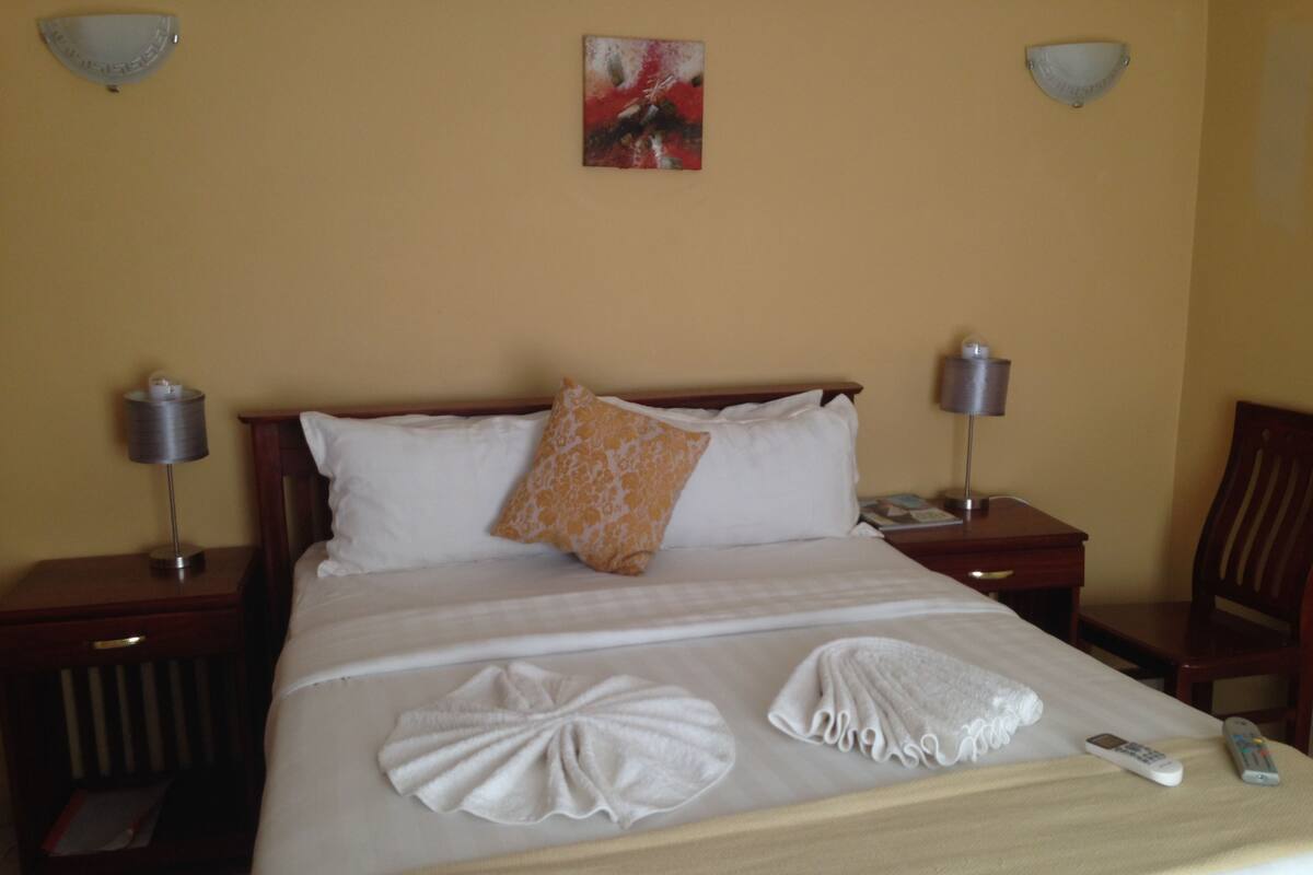 A neatly made bed with white linens, two pillows, and decorative towels. There are two bedside tables with lamps, a painting on the wall, and a chair to the side.