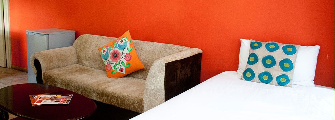 Una habitación con un sofá y una cama, ambos con almohadones decorativos. Hay una mesita con una revista encima.