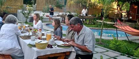 Un groupe de personnes est assis à une table en plein air et prend son repas, avec une piscine en arrière-plan.