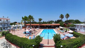 A swimming pool surrounded by lounge chairs and umbrellas, with palm trees and a building in the background. - Sandy's Studios Apartments & Log Villas (Kos)