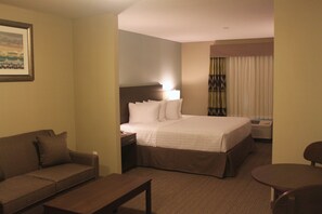 A hotel room with a bed, a bedside table, a lamp, a sofa, a small round table, and a window with curtains. - Meridian INN Suites (Lloydminster)