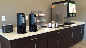 Una estación de café autoservicio con varias cafeteras, un dispensador de bebidas y un refrigerador con pantalla digital.