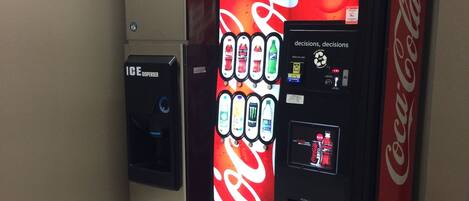 A Coca-Cola vending machine and an ice dispenser in a room with beige walls and tiled flooring.