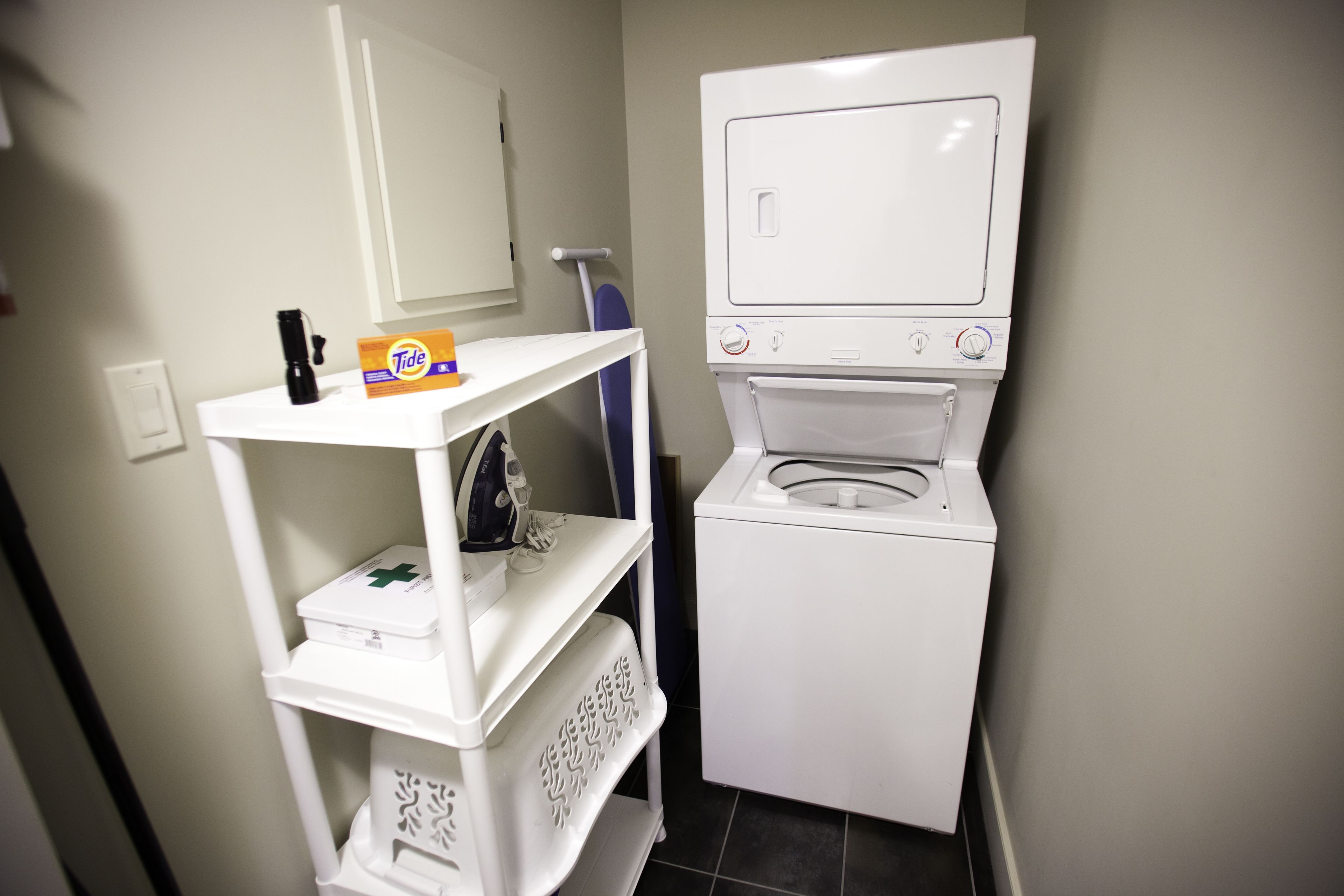 A compact laundry area with a front-loading washing machine, a shelf with a box of Tide detergent, and a small ironing board.