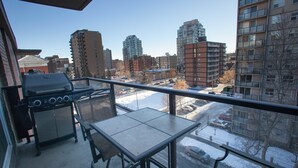 A balcony with a grill, table, and chairs, overlooking a snowy street and buildings. - Corporate Suites of Calgary - Eightwelve (Calgary)