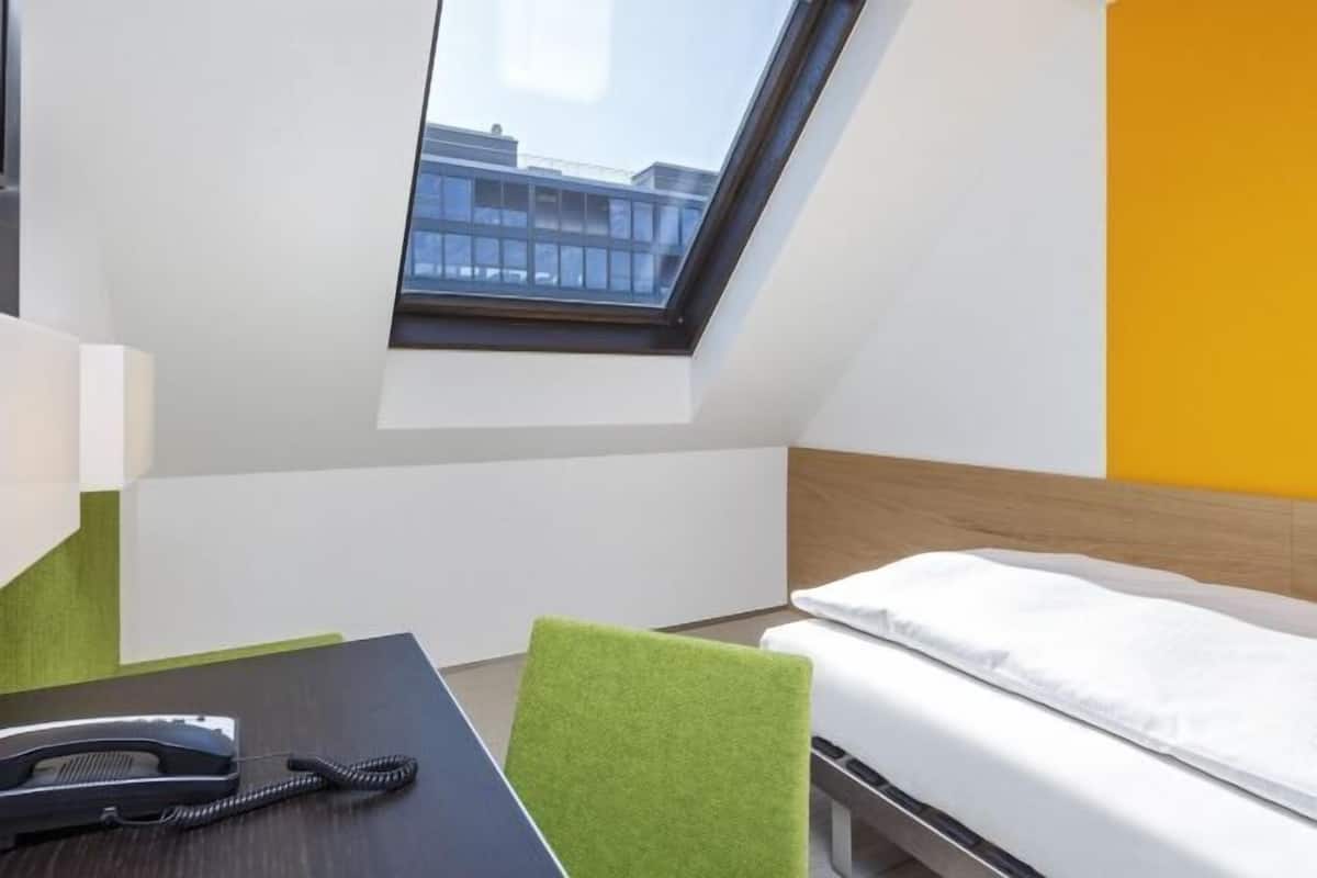 Quarto de hotel com clarabóia, uma cama, uma escrivaninha com telefone e uma cadeira verde.