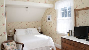 A bedroom with floral wallpaper, a bed with white bedding, a wooden nightstand with a lamp, a dresser, and a television on top.