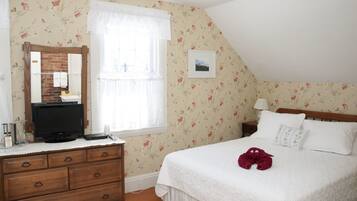 A bedroom with a wooden dresser, a television, a window with a white curtain, a bed with white bedding, and a red towel on the bed.