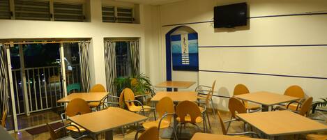 Un área de comedor con mesas y sillas, un televisor montado en la pared y una ventana con cortinas.
