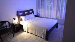 A neatly made bed with a wooden headboard, a bedside table with a lamp, and a chair. - Bayfront Hotel (Fort-de-France)