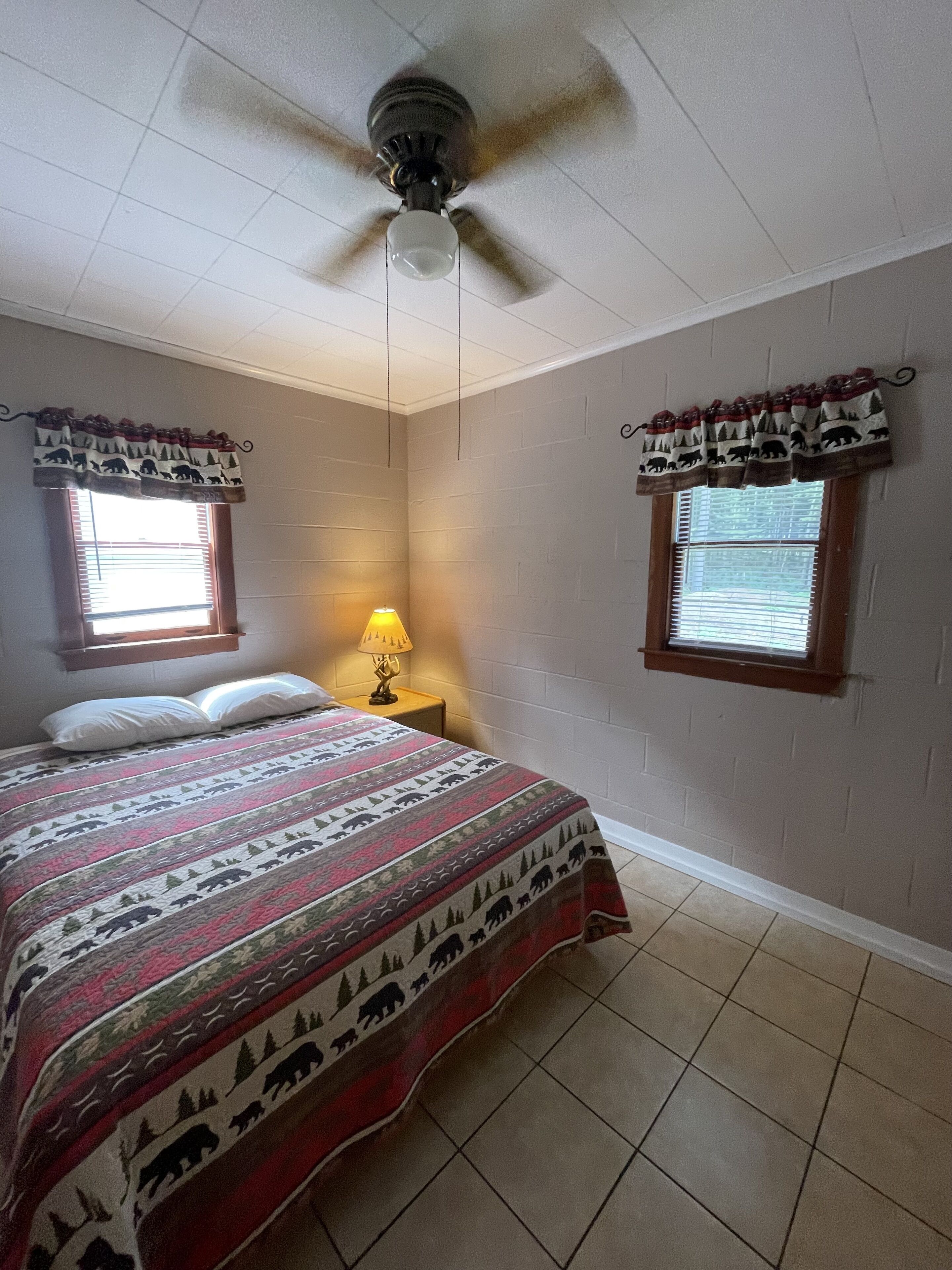 A bedroom with a bed, two windows with curtains, a ceiling fan, and a nightstand with a lamp.