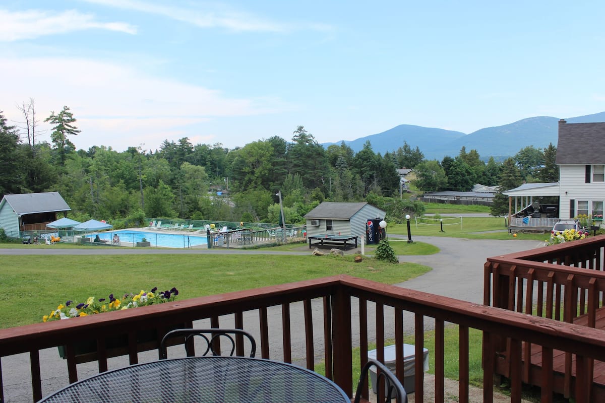 A deck with a metal table and chairs, overlooking a swimming pool and a small building with a Pepsi sign.