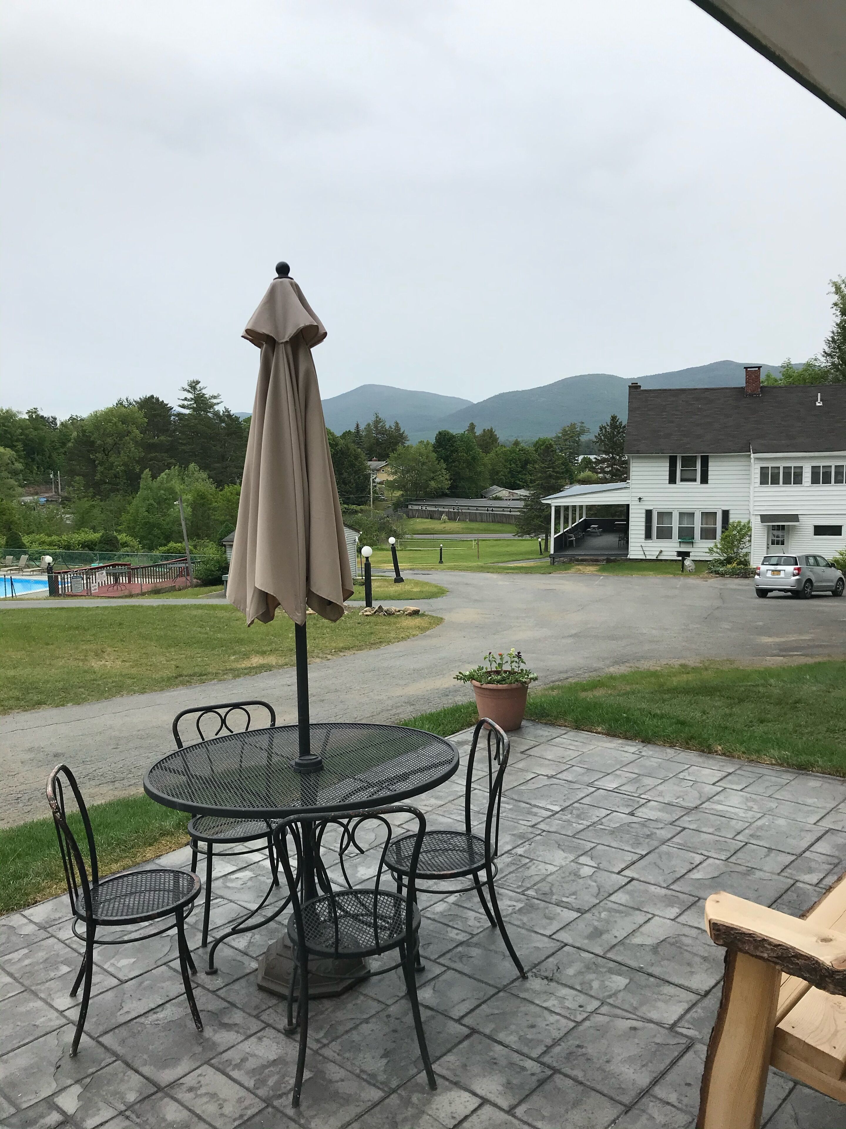 A patio with a round table and chairs, a beige umbrella, and a view of a house and mountains in the background.