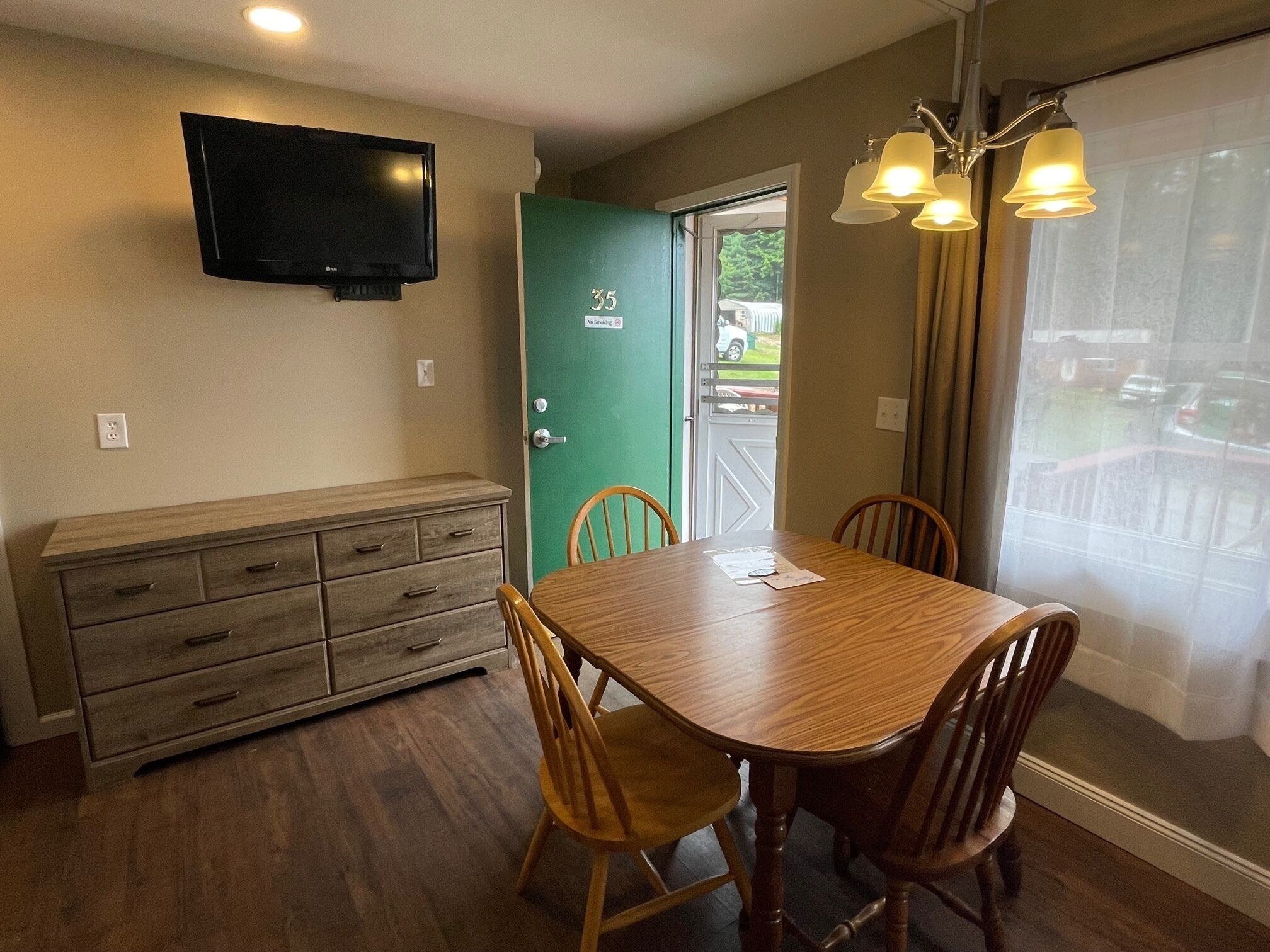 A dining area with a wooden table and chairs, a television mounted on the wall, and a green door with a 'No Smoking' sign.