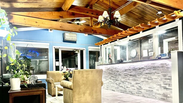 A spacious room with a high ceiling, wooden beams, and a chandelier. There are two beige armchairs and a small table with a potted plant. A large window provides a view of the outside. - Crossroads Motel (Elkhorn)