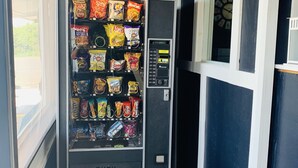 A vending machine with various snacks and drinks, located in a hallway with a clock and a window. - Crossroads Motel (Elkhorn)